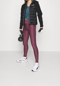 Chaqueta puffer negra sobre una blusa verde azulado, combinada con leggings texturizados en color marrón y zapatillas blancas con acentos en rosa y verde. Sostenida en la mano, una pesa rusa negra.