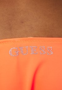 Oranžno kopalno oblačilo z kristalno dekoracijo, ki na sprednji strani črkuje "GUESS". Ima gladko teksturo in strukturirano zasnovo.