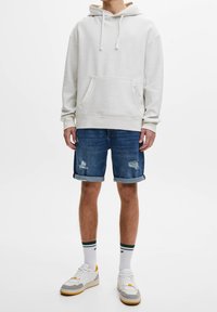 Felpa grigia chiaro con cappuccio, in misto cotone, tasca a marsupio. Pantaloni corti in denim blu con dettagli strappati, polsini arrotolati. Sneakers bianche con dettagli colorati.
