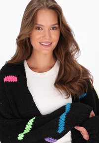 Melna adīta kardigāna ar daudzkrāsainiem zīmogojuma rakstiem rozā, zaļā, zilā un purpursarkanā krāsā. Uzvilkta virs baltas garām piedurknēm.