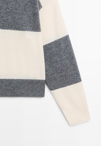 Manica di maglione a righe orizzontali larghe in tessuto lavorato a maglia grigio e bianco sporco, con dettaglio di polsino e orlo a costine su sfondo bianco.