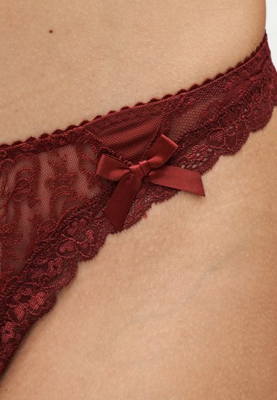 Gros plan sur la hanche d'une personne portant une culotte en dentelle bordeaux avec un petit nœud en satin sur un tissu transparent.