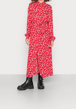 Kobieta w czerwonej sukience z długim rękawem z białymi nadrukami stokrotek, z paskiem wiązanym w talii i czarnymi masywnymi botkami, stojąca na szarej podłodze.