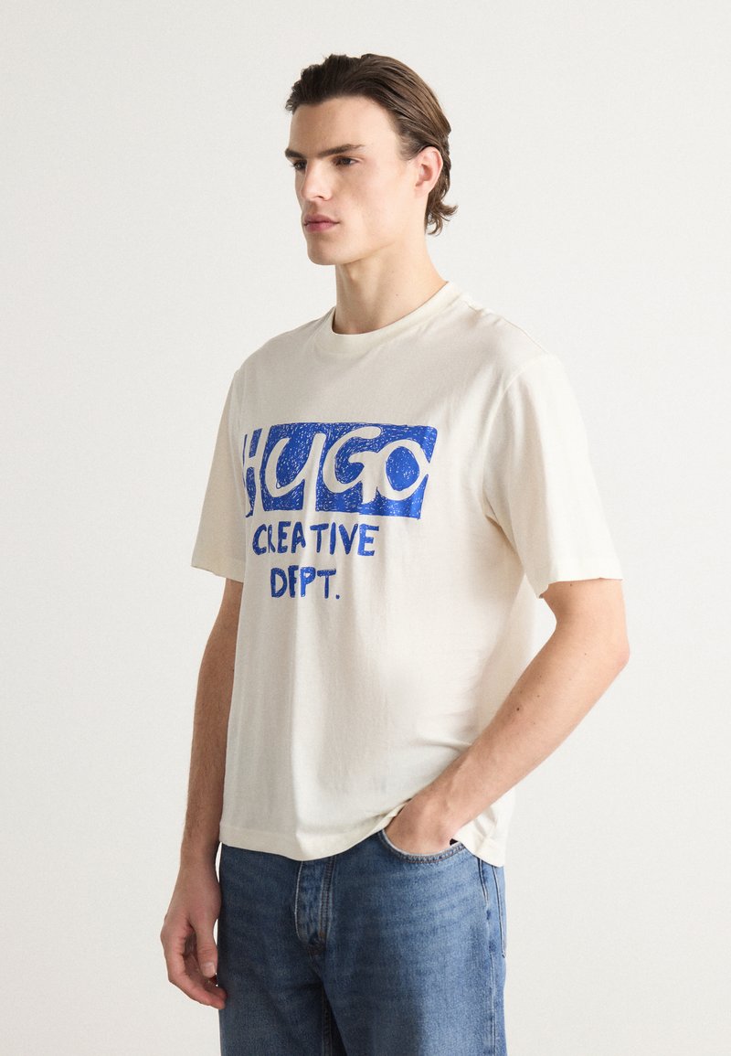 Kremfarget bomulls t-skjorte med korte ermer, med blå tekst "HUGO CREATIVE DEPT." i fet, teksturert trykk.