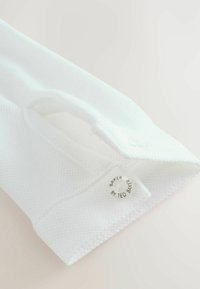 Polo bianca in cotone con motivo testurizzato, con un bottone etichettato "BAKER BY TED BAKER" vicino al colletto. Finitura liscia, design piegato.