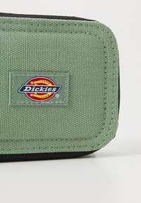 Zaļa auduma maks ar melniem akcentiem, taisnstūrveida formā. Iekļauts izšūts Dickies logo uzlīme priekšpusē.