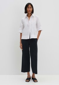 Camicetta bianca a maniche corte con colletto a punta, abbinata a pantaloni scuri larghi e sandali neri a papera. Design semplice e minimalista.