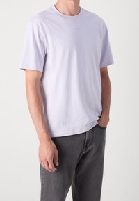 Mężczyzna w jasnofioletowej koszulce z krótkim rękawem i ciemnoszarych dżinsach, z jedną ręką w kieszeni, a drugą swobodnie wzdłuż ciała.