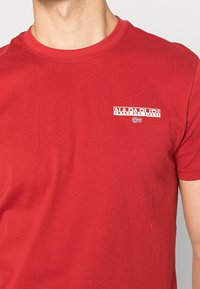 Rød bomulds t-shirt med rund hals, korte ærmer, og et hvidt "NAPAPIJRI" logo med et lille flag-grafik på forsiden.