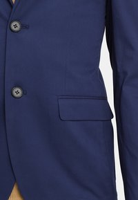 Mörkblå blazer med två svarta knappar och en ficka med flik på höger sida, baren över en beige tröja.