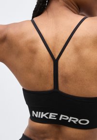 Γυναίκα που φοράει μαύρο αθλητικό σουτιέν Nike Pro με λεπτές λωρίδες σε σχήμα Υ στην πλάτη, εμφανιζόμενη από πίσω πάνω σε ουδέτερο φόντο.