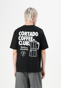 Schwarz Baumwoll-T-Shirt mit weißem Text und einem Grafikmotiv eines Kaffeeautomaten auf dem Rücken. Kurze Ärmel, lockere Passform, einfaches Design.