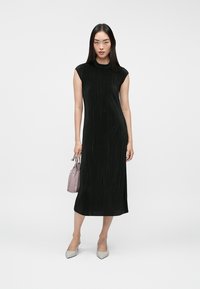 Nő fekete ujjatlan midi ruhát visel, egy kis mályvaszínű táskát tart a kezében, és világosszürke, hegyes orrú magassarkú cipőt visel.