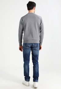 Šedá mikina s dlouhými rukávy, žebrovanými manžetami a lemem, v kombinaci s modrými džíny a bílými teniskami, pohled zezadu.