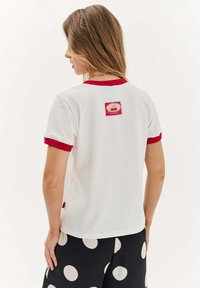 Bílý tričko s krátkým rukávem, červeným žebrovaným límcem a rukávy, s nášivkou na zadní straně s designem třešně a textem „NAOKO.“