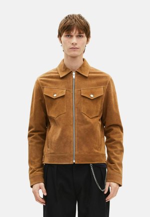 Giacca in suede marrone con colletto, cerniera frontale e due tasche sul petto. Presenta dettagli in argento e una catena decorativa.