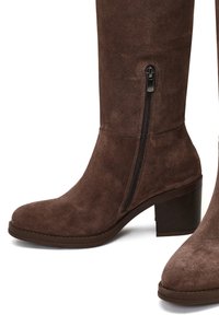 Bruine suede kniehoge laarzen hebben een ronde neus, een chunky hak en een zijrits. Het materiaal heeft een zachte textuur, wat het totale ontwerp versterkt.