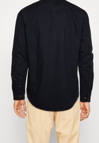 Chemise noire à manches longues avec un ourlet arrondi, des poignets à boutons et une texture lisse, associée à un pantalon beige clair.