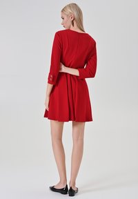 Rode jurk met lange mouwen, nauwsluitend lijfje, uitlopende rok, geruite manchetten en een ritssluiting aan de achterkant. Draagt zwarte lakvlats.