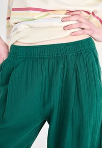 Groene geplooide broek van zachte stof, met een elastische tailleband en een gestructureerd oppervlak. De outfit wordt aangevuld met een gestreept topje.