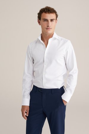 Camisa blanca de manga larga con cuello clásico y botones, combinada con pantalones ajustados en azul marino. Textura de tela suave, diseño de corte slim.