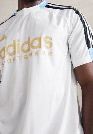 Hombre vistiendo una camiseta deportiva blanca de Adidas con logo dorado, ribete de cuello azul claro y rayas negras en las mangas.