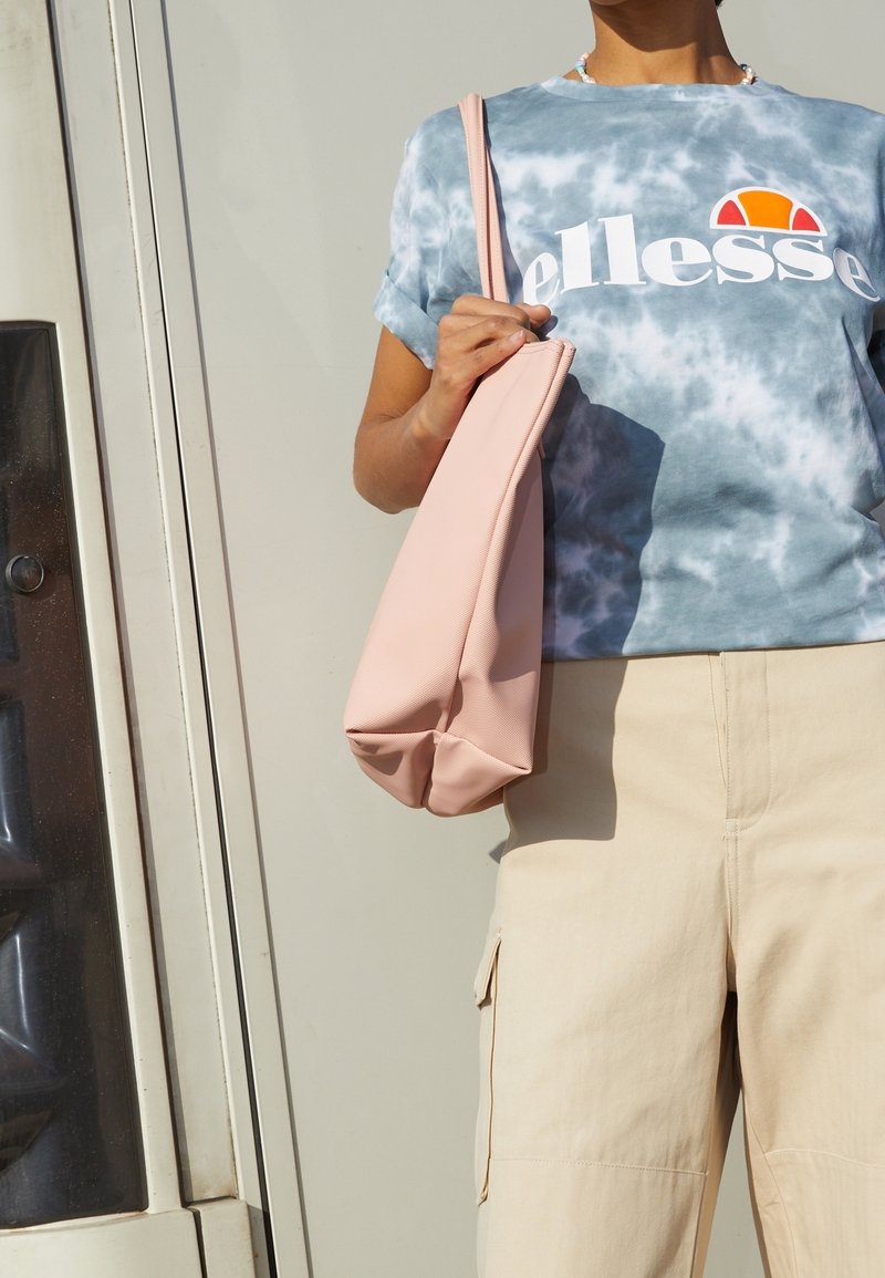 T-shirt bleu et blanc tie-dye avec un logo blanc, associé à un pantalon cargo beige clair. Tenue dans un sac structuré doux et rose.
