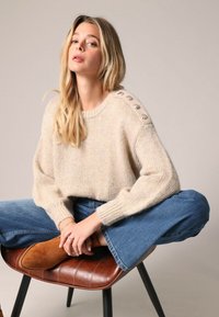 Pull en tricot beige avec de larges manches, col rond et grands boutons décoratifs sur une épaule ; associé à un jean bleu évasé et des chaussures marron.