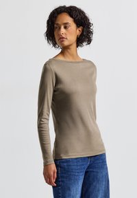Langarmshirt aus einem weichen, hellbraunen Stoff mit einem Boots-Ausschnitt, figurbetonter Form und glatter Textur. Kombiniert mit Jeans.