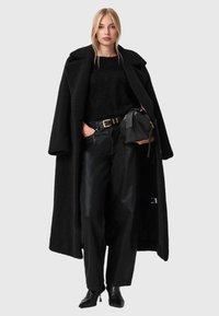 Černý strukturovaný kabát, černý chlupatý svetr, lesklé černé kalhoty s širokými nohavicemi a černé boty na podpatku, držící malou černou kabelku.