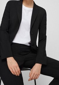 Svart skräddarsydd kavaj och byxor i ett mjukt material, i kombination med en vit t-shirt med rund hals. Kavajen har två knappar.