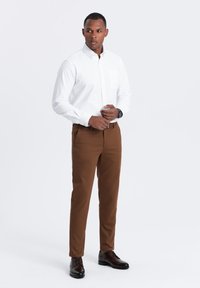 Weißes Button-Down-Hemd, braune Hose und dunkle Schuhe. Das Hemd hat eine Brusttasche. Die Hose hat eine strukturierte Oberfläche und ist tailliert.