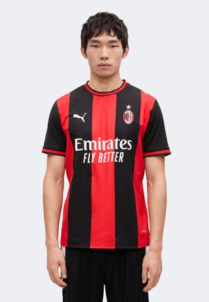 Ung mand står iført en AC Milan fodboldtrøje med røde og sorte lodrette striber og Emirates Fly Better-logo.