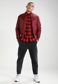 Giacca bomber bordeaux, camicia a quadri rossa e nera sotto, pantaloni grigio scuro e sneakers bianche. Modello in piedi con le mani in tasca.