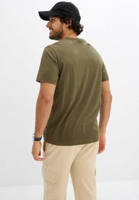 T-shirt in cotone verde oliva con maniche corte e scollatura rotonda, abbinato a pantaloni cargo beige, indossato da una persona vista da dietro.