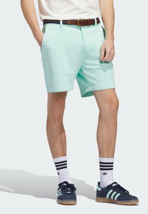 Person trägt helltürkise Shorts mit Seitentaschen, weiße Kniestrümpfe mit schwarzen Streifen, marineblaue Sneaker mit hellen Streifen und einen braunen Gürtel.