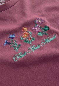 Tissu violet avec des fleurs brodées au point de croix en bleu, orange et rose, ainsi que des feuilles vertes, au-dessus d’un texte en écriture cursive indiquant « Fleur Après Minuit ».