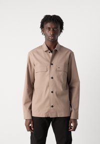 Beige syntetisk skjorte med knaplukning og to brystlommer, rund kant og sorte knapper. Lange ærmer med manchetter, båret over sorte bukser.