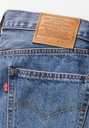 Denim jeans with a light blue wash, featuring a brown leather back patch, orange stitching, and a red logo tag on the left pocket.