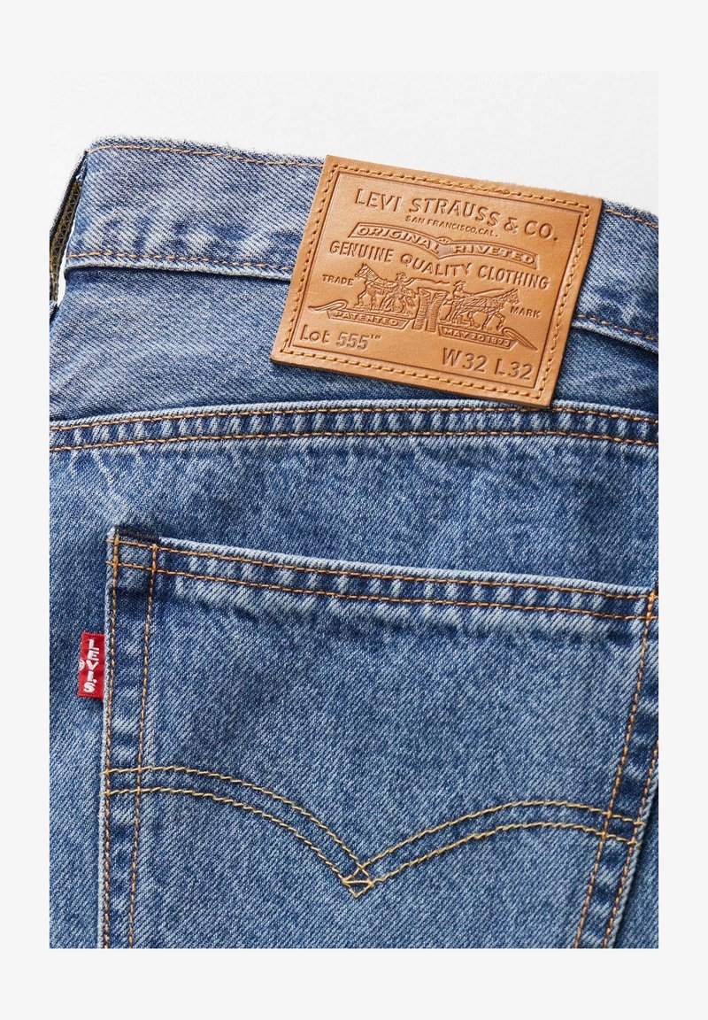 Vaqueros de mezclilla con un lavado azul claro, que cuentan con un parche trasero de cuero marrón, costuras naranjas y una etiqueta con logo roja en el bolsillo izquierdo.