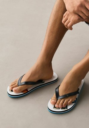 Barfuß in weißen Flip-Flops mit schwarzen Riemen, auf einem hellgrauen Boden ruhend, Hände über den Beinen verschränkt.