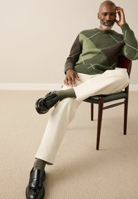 Green and brown patterned sweater, cream trousers, dark loafers, and olive socks. Seated on a wooden chair with a textured beige floor.