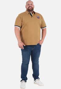 Polo marron avec des accents noirs et blancs, présentant un logo brodé. Porté avec un jean bleu et des baskets blanches. La personne se tient devant un fond blanc.