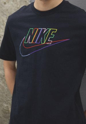 Personne portant un t-shirt noir avec un logo Nike multicolore et un swoosh en contour sur la poitrine, devant un fond texturé.