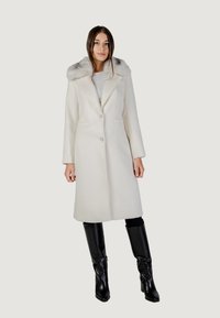 Manteau en laine blanche avec un col en fourrure gris, design croisé avec double boutonnage, poches latérales et coupe ajustée, associé à des bottes noires montantes jusqu'aux genoux.