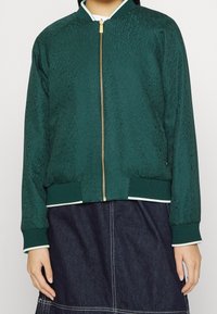 Zelená bomber bunda z texturované látky se zlatým zipem. Má žebrované manžety a lem s bílými akcentními pruhy. Nasažena přes modrý top.