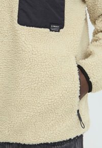 Fleece pullover in cream with a textured surface, featuring a black chest pocket and a side zipper. Label reads "11 PROJECT."
