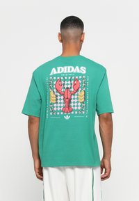 Türkiz színű pamut póló, rózsaszín homárral, kockás mintával, és "ADIDAS ORIGINALS" felirattal a hátsó részén. Rövid ujjú.