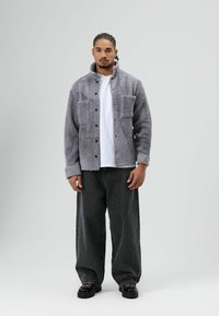 Grijze fleecejas met twee voorzakken, knoopsluiting en een zachte textuur, gecombineerd met zwarte wijd uitlopende jeans en zwarte schoenen.