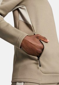 Top deportivo beige con una textura suave, paneles laterales en contraste y un bolsillo con cremallera en la parte delantera derecha.